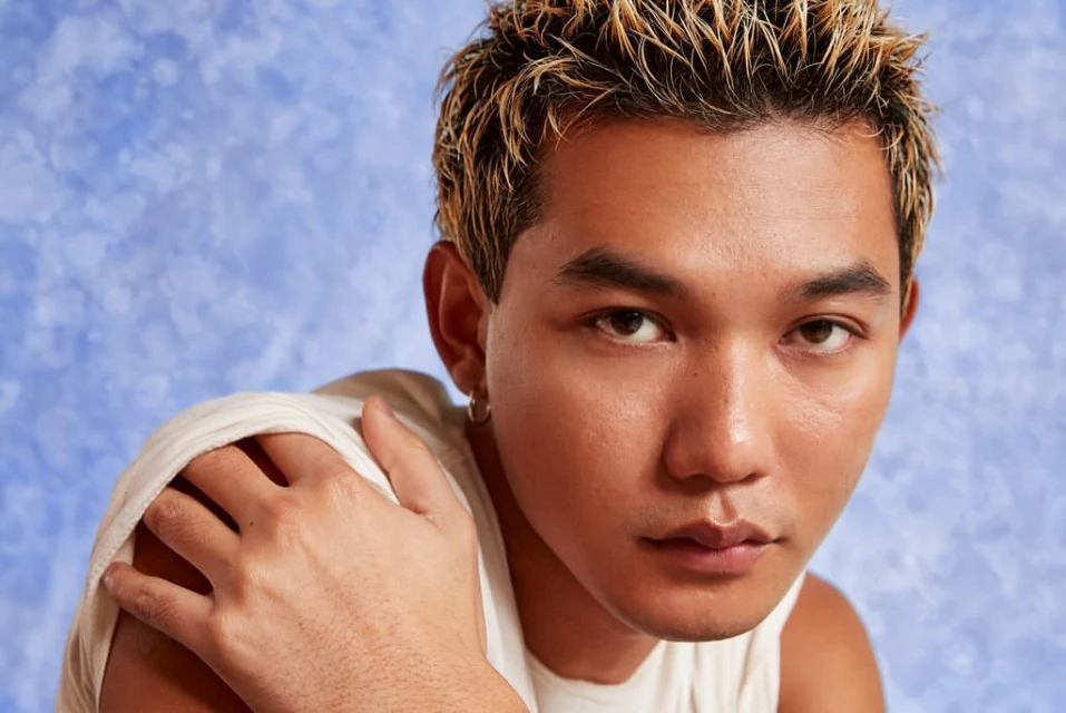 Close-up portrait of a tan-skinned adult male with blond-streaked hair, wearing a white tank top, against a blue mottled background.