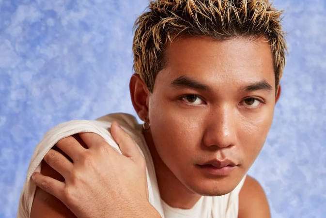 Close-up portrait of a tan-skinned adult male with blond-streaked hair, wearing a white tank top, against a blue mottled background.