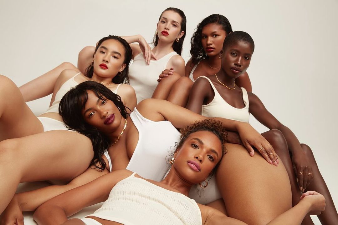A group of diverse women posing together in white tops, showcasing natural beauty and minimalistic fashion against a neutral backdrop.