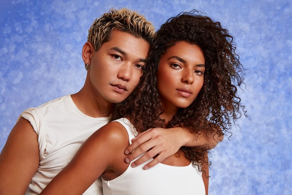 Portrait of an Asian man and a biracial woman looking at the camera. The couple is wearing white tops against a blue backdrop.
