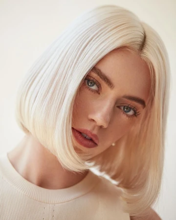 Close-up portrait of a woman with platinum blonde bob hair and blue eyes wearing a cream top against a neutral background