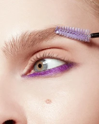 Extreme close-up of a blue eye with purple eyeliner being brushed with a spoolie brush by photographer Beth Sternbaum.