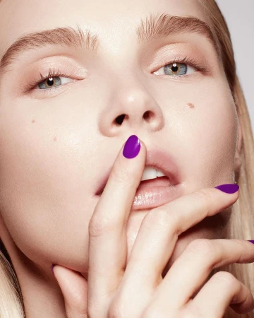 Close-up beauty portrait of a woman with vibrant purple nail polish touching her lips against a neutral background.