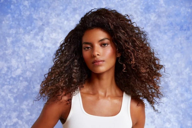 Close-up portrait of a woman with voluminous curly dark hair wearing a white tank top against a mottled blue background.