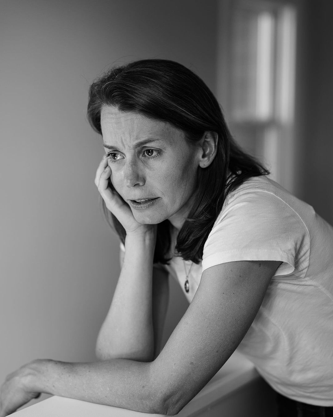 Monochrome portrait of a pensive woman with dark hair, resting her head on her hand indoors, with a diffused light source.
