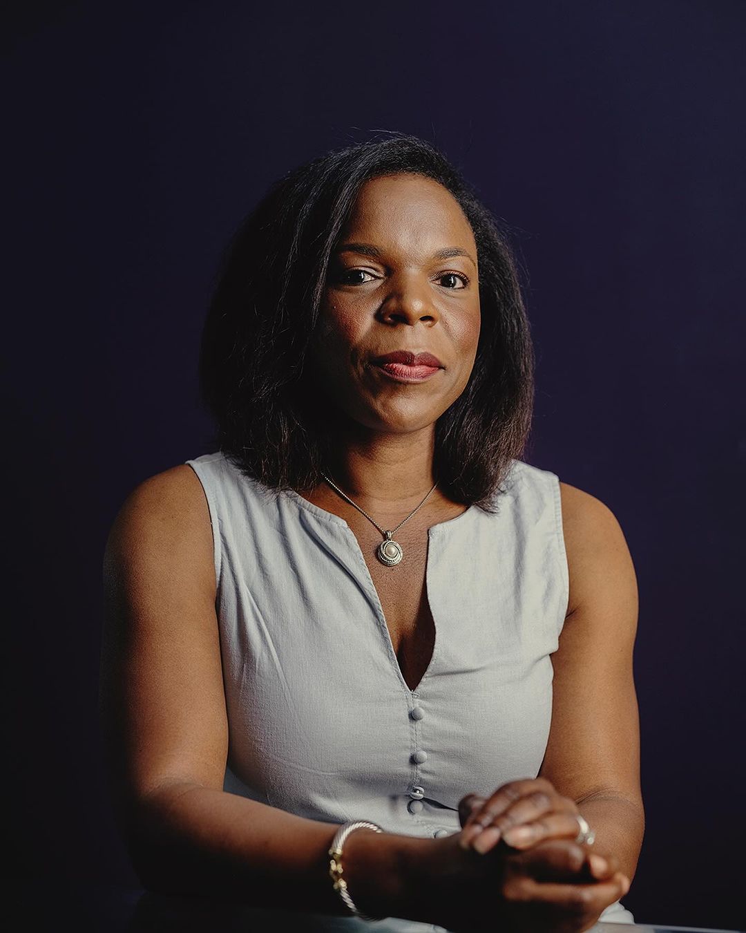 Portrait of a black woman with dark hair wearing a sleeveless dress, a necklace, and bracelets against a dark blue background.