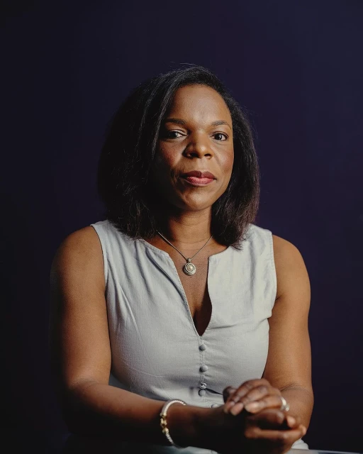 Portrait of a black woman with dark hair wearing a sleeveless dress, a necklace, and bracelets against a dark blue background.