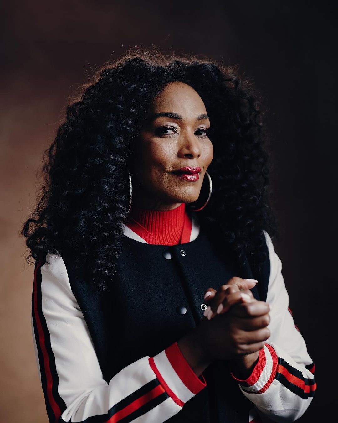 Close-up portrait of a woman with voluminous curly hair, wearing a red turtleneck sweater and a bomber jacket against a brown backdrop.