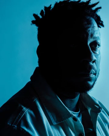 Portrait of a Black man with short dreadlocks wearing a light collared shirt against a blue background, shot by Bethany Mollenkof.