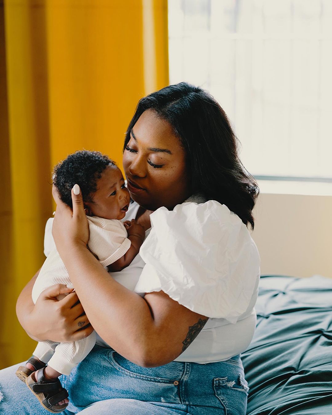 An African-American mother holds her newborn baby indoors, with soft window light and a yellow accent wall creating a cozy, loving scene.