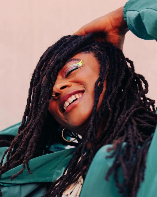 Smiling African-American woman with dreadlocks and creative makeup posing in front of pink wall wearing green jacket, close-up.