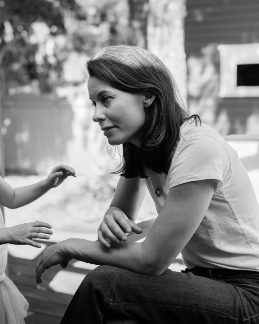 Black and white portrait of a woman sitting with a young child outdoors in a backyard setting, interacting with the child's hands.
