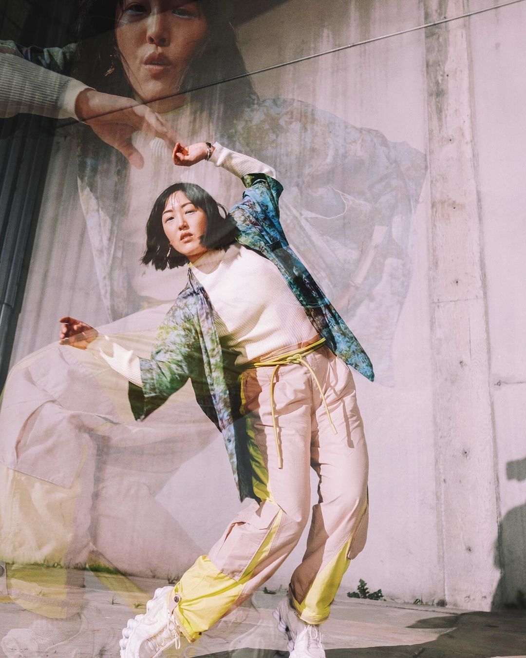 Asian woman in beige pants and jacket, stands in a dynamic pose outdoors, with a translucent double exposure effect in background.