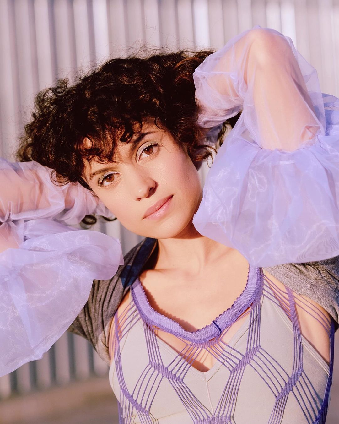 Close-up portrait of a woman with curly hair wearing a unique lavender top and sheer sleeves, looking directly at the camera in a bright, airy setting.