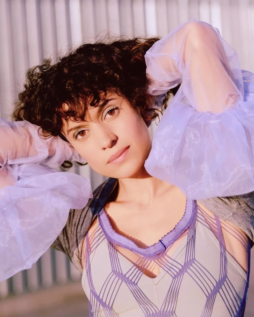 Close-up portrait of a woman with curly hair wearing a unique lavender top and sheer sleeves, looking directly at the camera in a bright, airy setting.