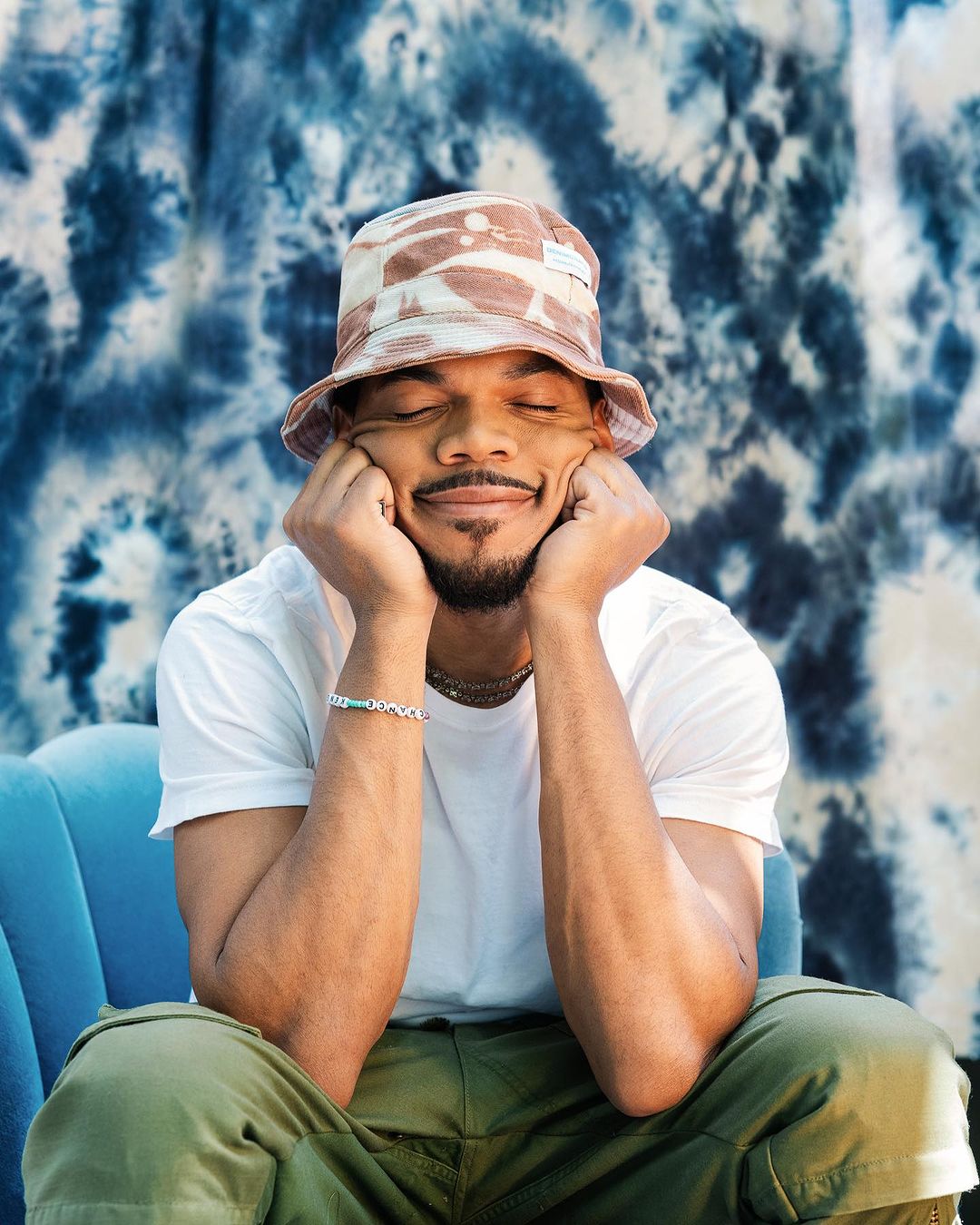 Man with a goatee poses with closed eyes, wearing a bucket hat, t-shirt and green pants, seated against a textured backdrop and blue sofa.