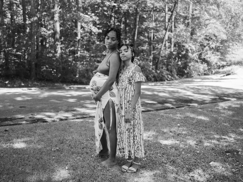 Pregnant woman with her daughter in a grassy field surrounded by trees. She wears a patterned dress, and the daughter wears a sundress.