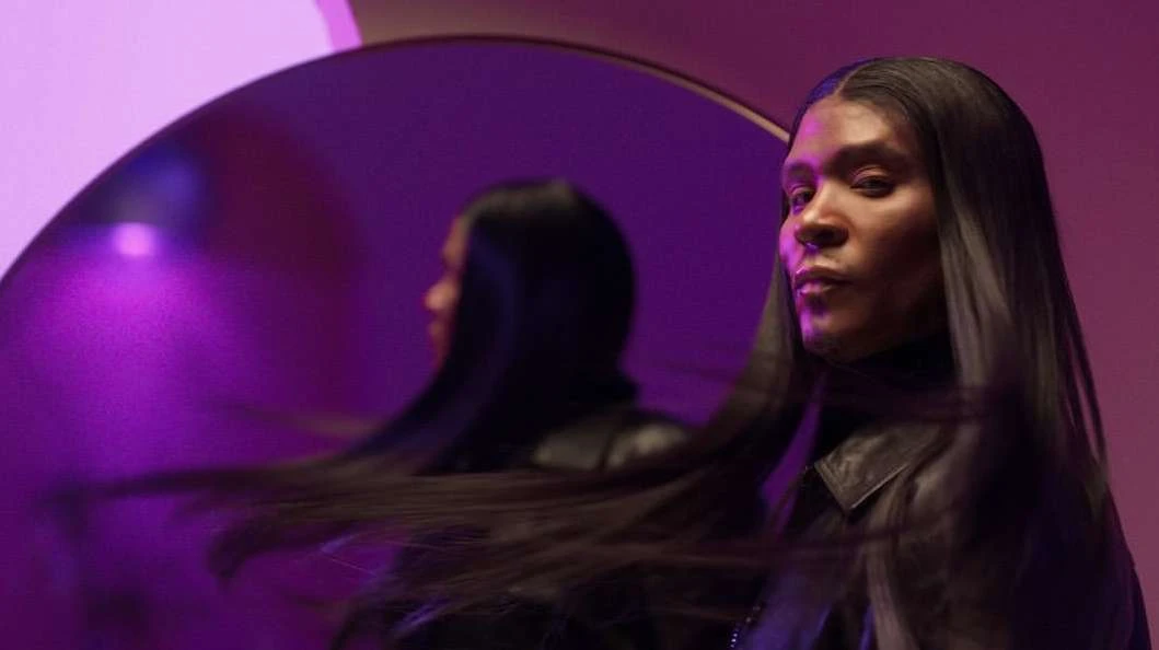 Person with long dark hair posing in front of a round mirror with a purple background and wearing a black leather jacket.