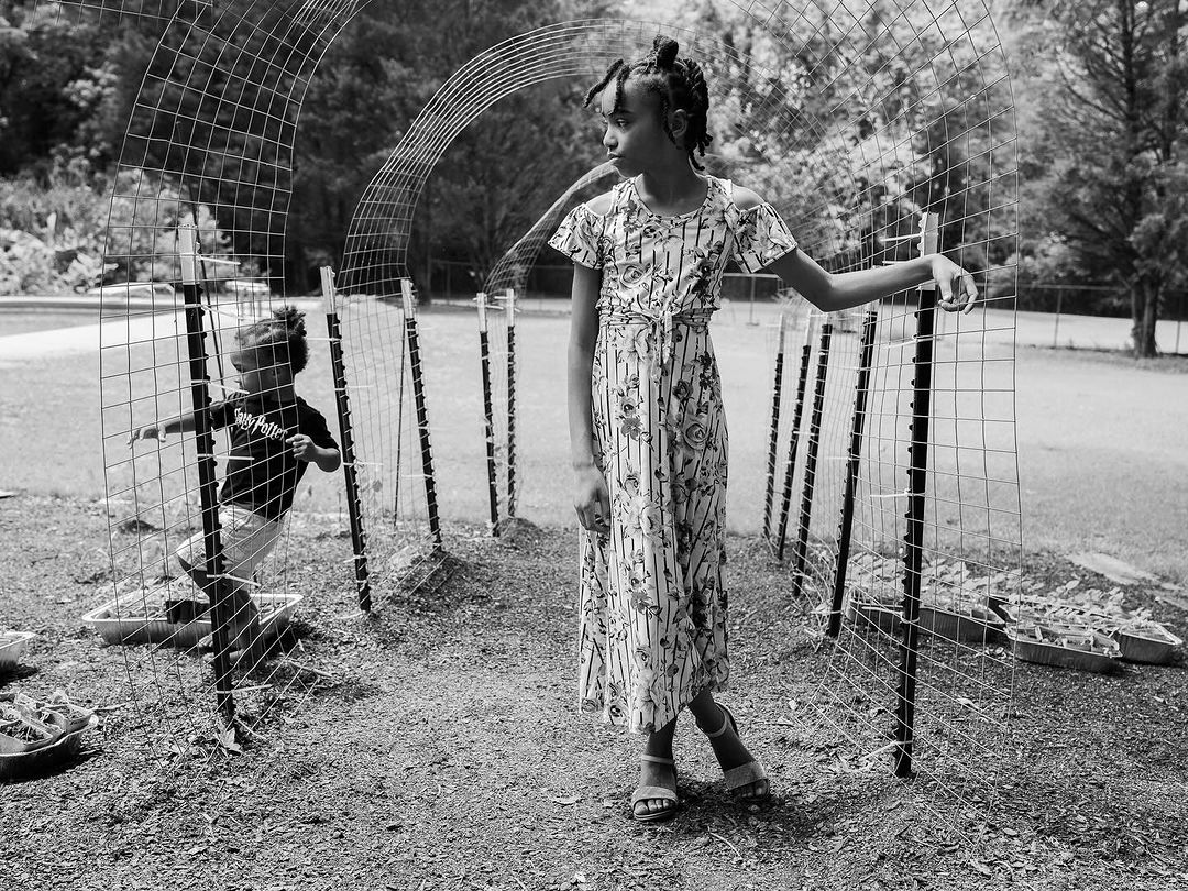 Two young African-American girls pose by a metal garden trellis on a sunny day. One wears a floral dress, the other a t-shirt.