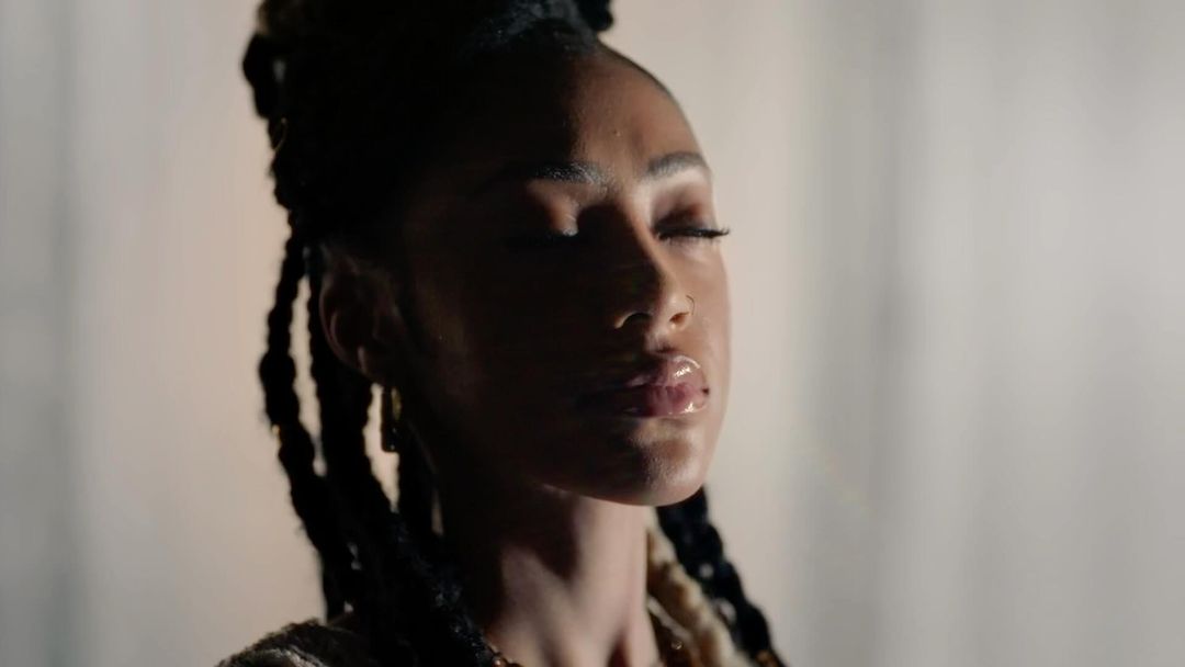 Portrait of a Black woman with braided hair and closed eyes, illuminated by dramatic side lighting against a neutral textured backdrop.