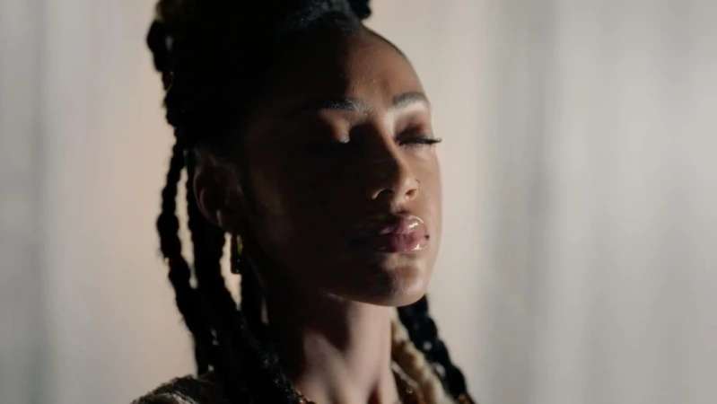 Portrait of a Black woman with braided hair and closed eyes, illuminated by dramatic side lighting against a neutral textured backdrop.