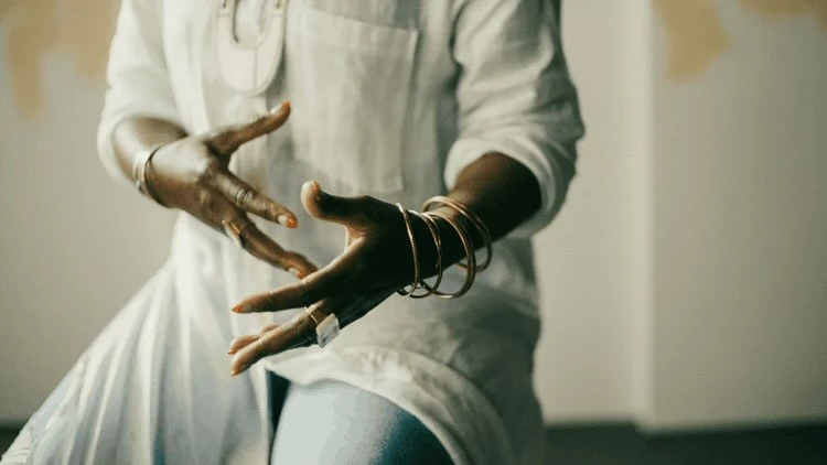Midsection view of an adult with arms raised, gold bracelets on the wrist, ring on the finger, and a white shirt inside a room.