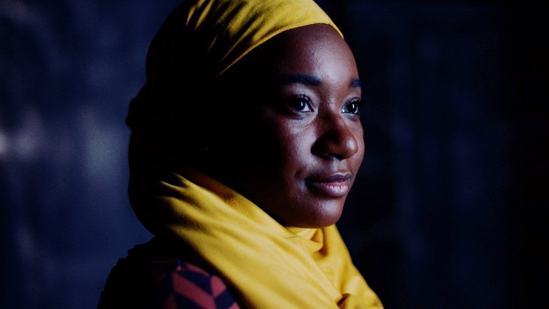 Headshot of an African American woman wearing a yellow headscarf. Low-key lighting enhances her features against a dark background.