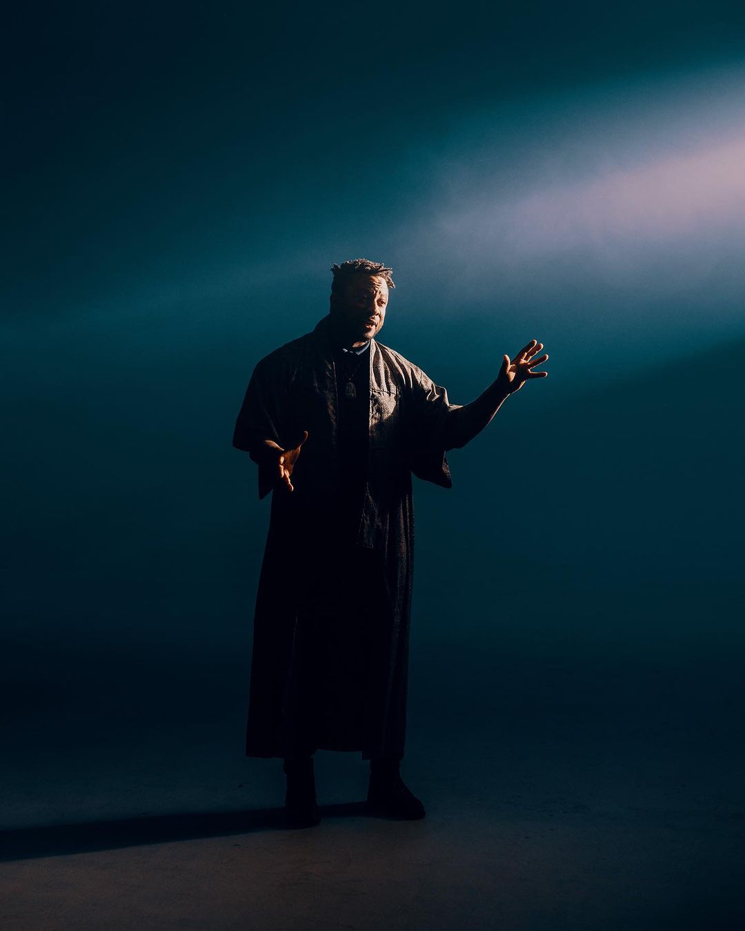 Man in dark robe stands with outstretched hands in front of a spotlight, wearing dark clothes against a shadowy blue background.