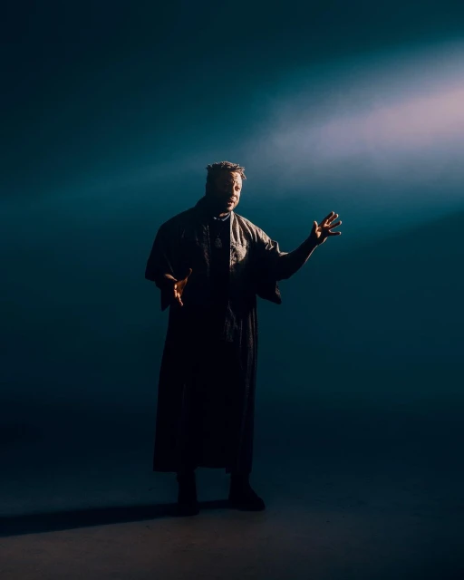 Man in dark robe stands with outstretched hands in front of a spotlight, wearing dark clothes against a shadowy blue background.