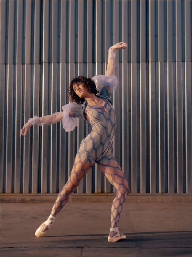 An adult female ballet dancer poses in front of a corrugated metal wall. She wears a patterned bodysuit and pointe shoes, with outstretched arms.