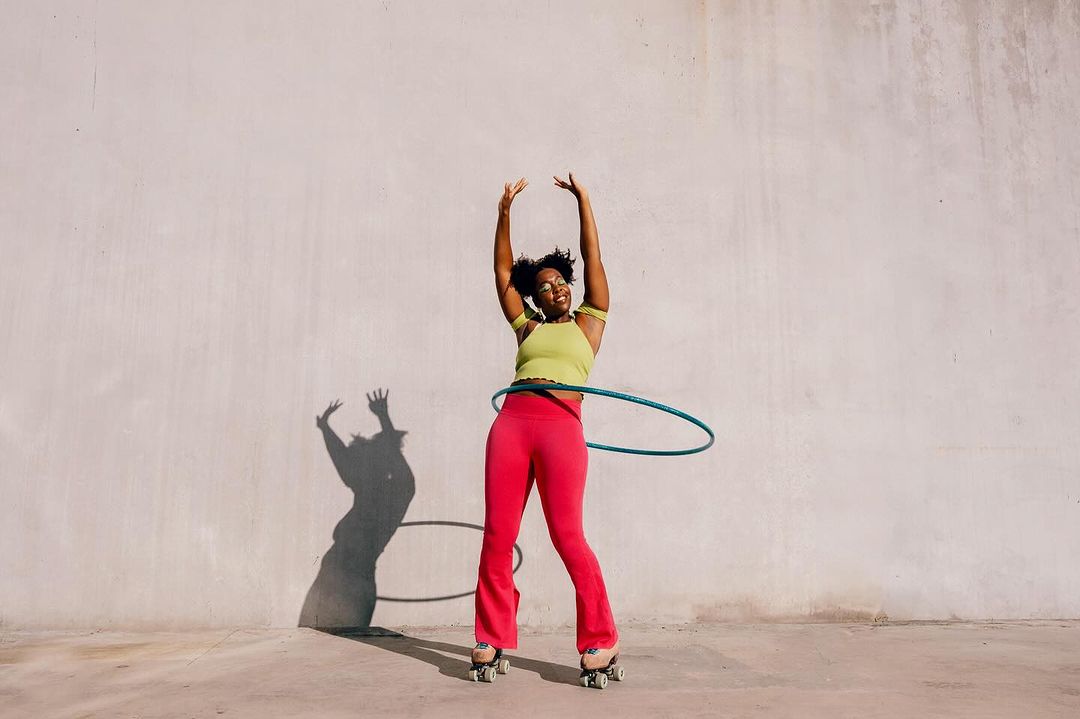 An adult woman with dark hair and glasses roller skates while hula hooping against a pale concrete wall, casting a shadow.