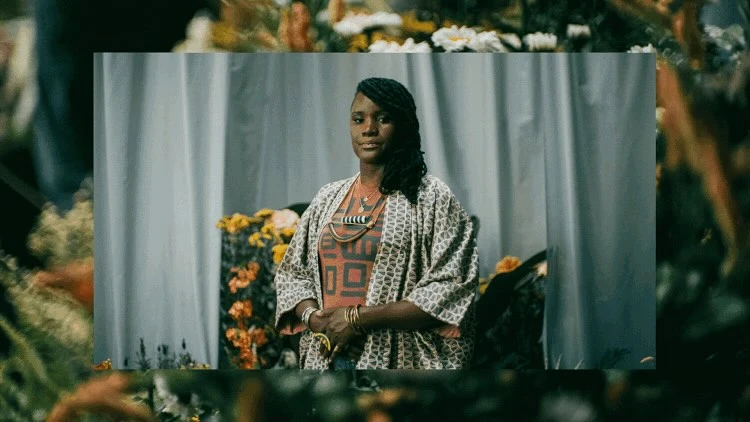 Portrait of an adult woman with dark hair, wearing ethnic jewelry and a patterned kimono-style robe, posing in front of white curtains.