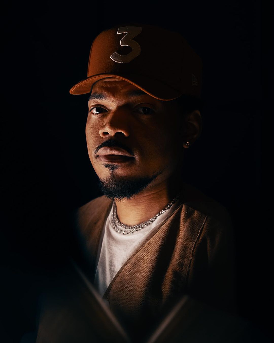 Portrait of a man wearing a brown baseball cap with a silver "3" on it and a white t-shirt against a dark backdrop.