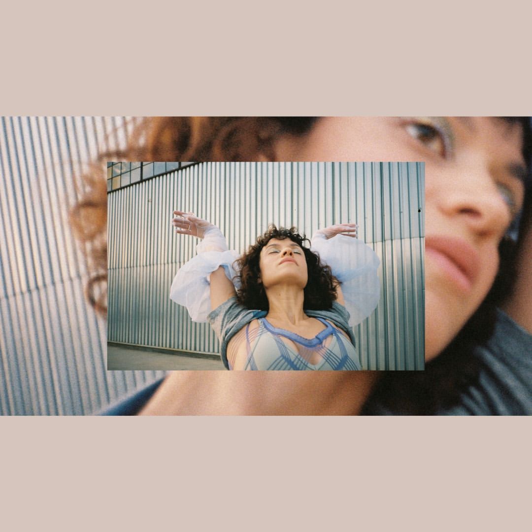 Portrait of a woman with arms raised, wearing a flowing top, against a textured metal wall, framed by a soft, out-of-focus overlay.