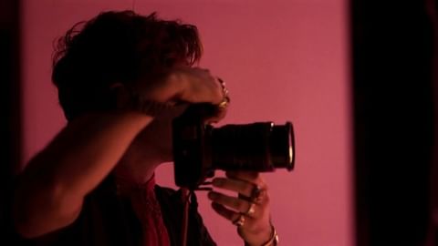 Person photographing with DSLR camera in dim, indoor lighting against a pink background; portrait of photographer in action.