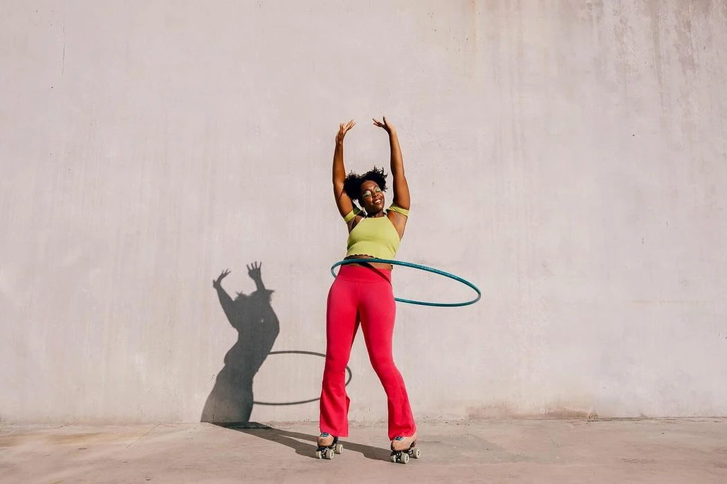 Black woman wearing pink pants and roller skates hula hooping against a plain concrete wall in New York