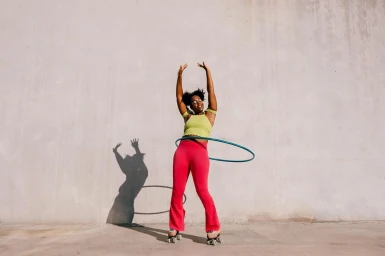 Black woman wearing pink pants and roller skates hula hooping against a plain concrete wall in New York