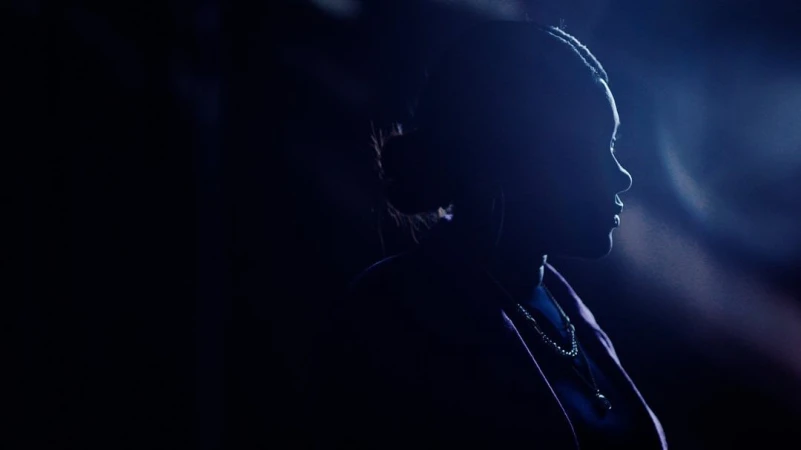 Silhouette of a Black woman in profile against a dark blue background with rim lighting on her hair and shoulder.