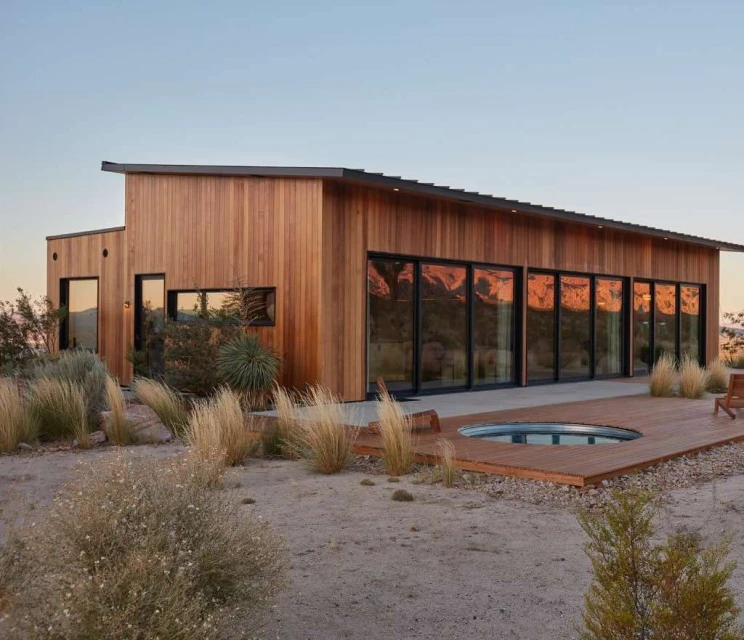 Exterior of a modern house with wood siding, large windows reflecting the desert landscape, and a built-in pool on a wooden deck.