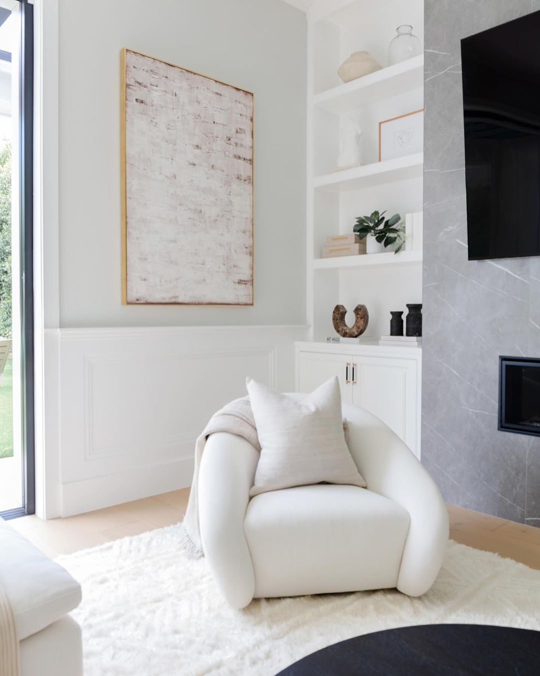 Cozy living room corner with a white armchair, decorative art, built-in shelves, a fireplace, and a soft rug.