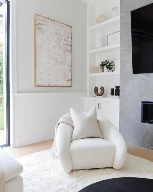Cozy living room corner with a white armchair, decorative art, built-in shelves, a fireplace, and a soft rug.