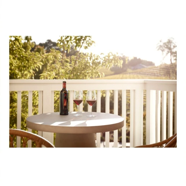 Wine bottle and two glasses of red wine on a table overlooking a vineyard with green trees under bright sunlight on a clear day.