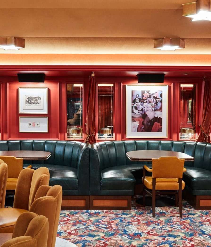 Interior of a restaurant with green booths, chairs, tables, red walls adorned with artwork, and a colorful patterned carpet.