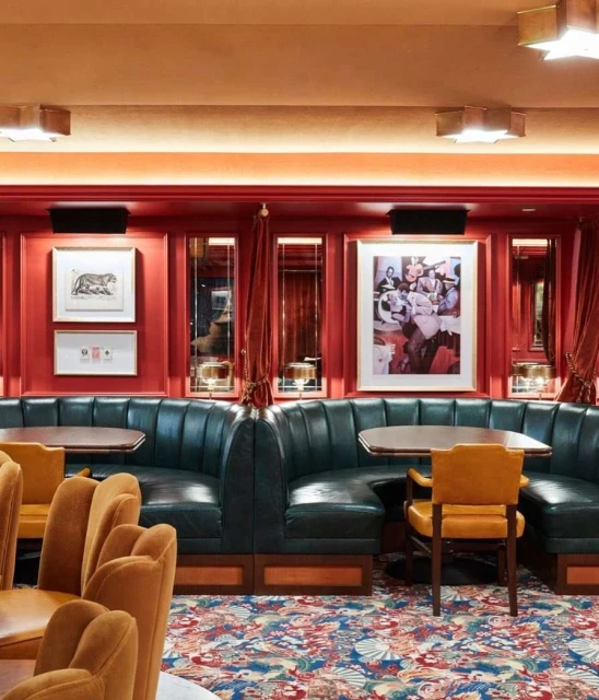 Interior of a restaurant with green booths, chairs, tables, red walls adorned with artwork, and a colorful patterned carpet.