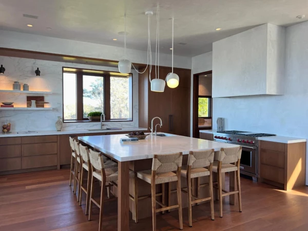 Brightly lit contemporary kitchen interior with a large island with multiple bar stools, wooden cabinetry, hardwood floors, and pendant lighting.