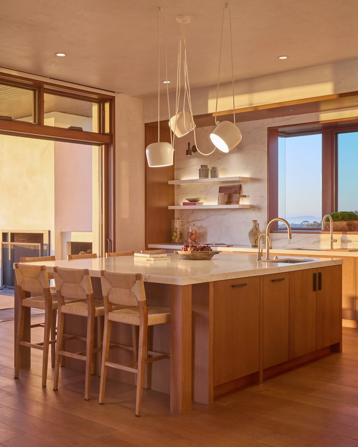Modern kitchen interior with a central island, pendant lights, and bar stools, illuminated by natural light and warm interior lighting.