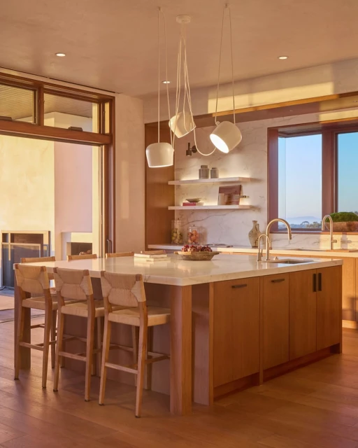 Modern kitchen interior with a central island, pendant lights, and bar stools, illuminated by natural light and warm interior lighting.
