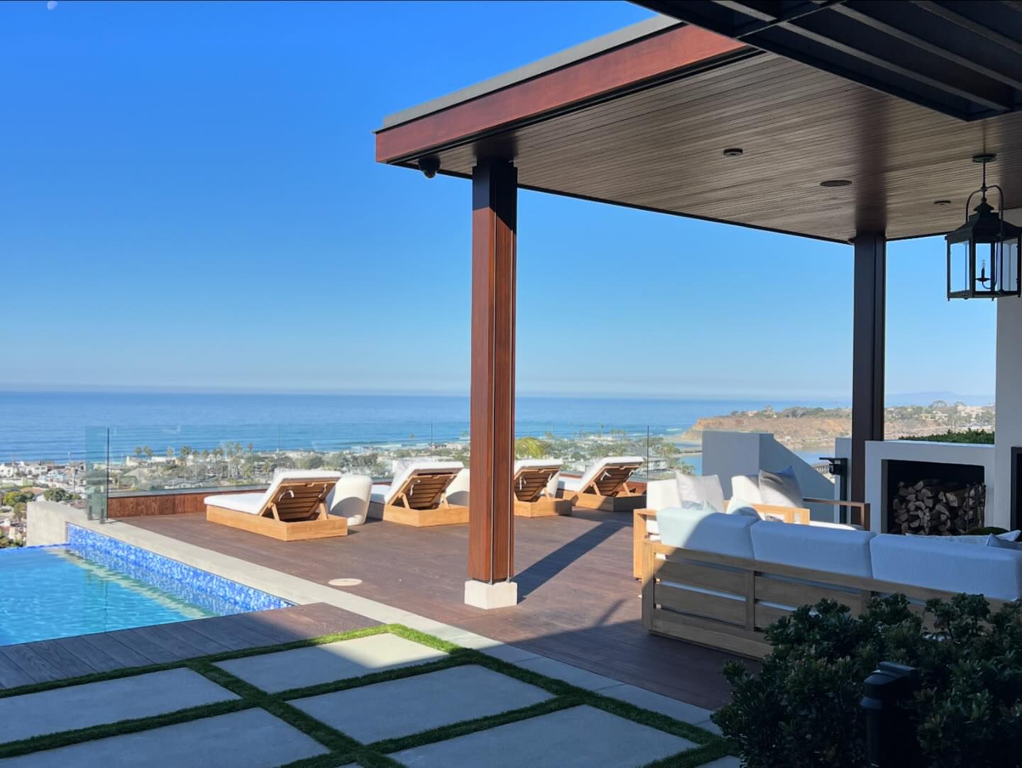 Outdoor patio with modern lounge furniture next to a blue swimming pool overlooking a coastal view of ocean and city on a sunny day.