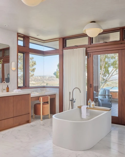 Bright bathroom interior featuring a white freestanding tub, vanity with a sink, stool, large window with a mountain view, and outdoor patio.