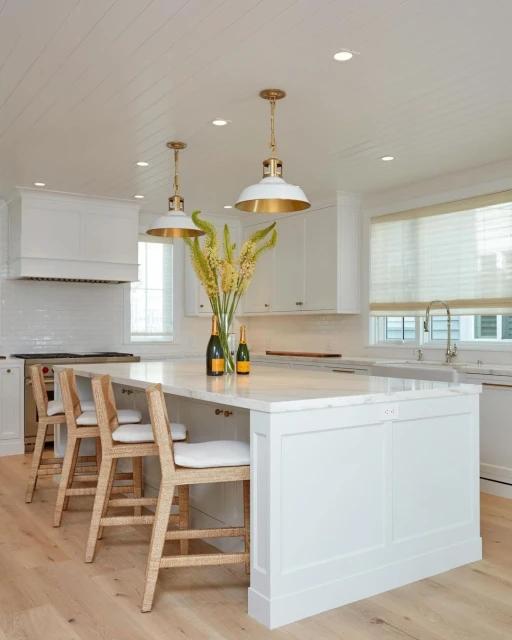 Bright, white kitchen interior featuring an island with bar stools, pendant lights, vase with floral arrangement and countertop items.